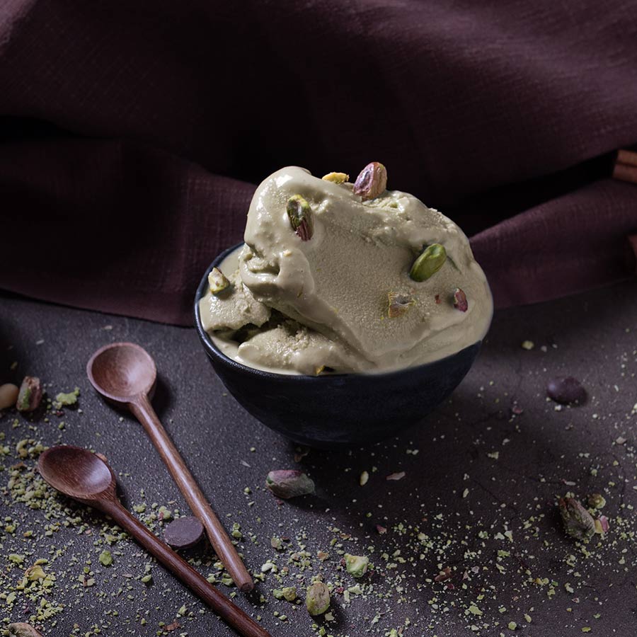 Gelato al pistacchio guarnito con pezzi di cioccolato e pistacchi interi, servito in una coppa bianca.