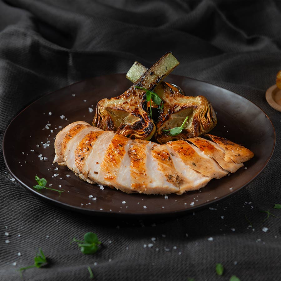 Petto di pollo grigliato con carciofi e prezzemolo su un piatto nero.