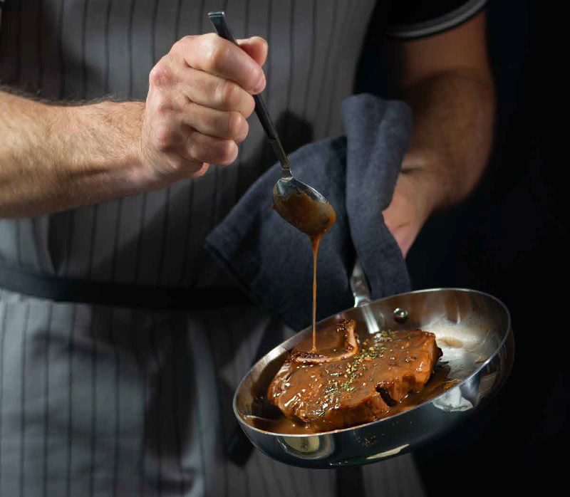 Chef in grembiule da cucina che versa salsa su una bistecca, mostrando abilità culinarie e attenzione ai dettagli.